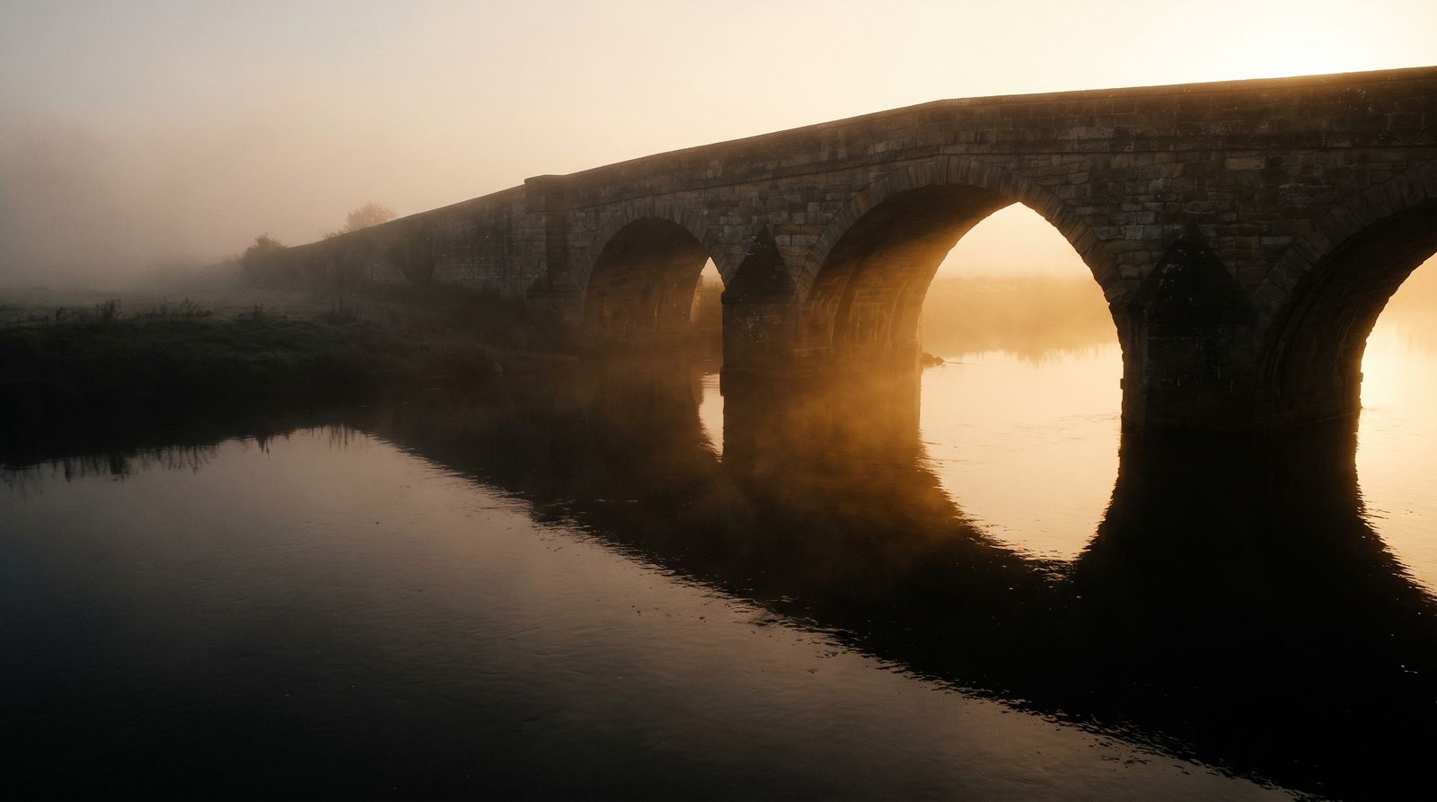 A stone arch bridge over a river at dawn, in mist